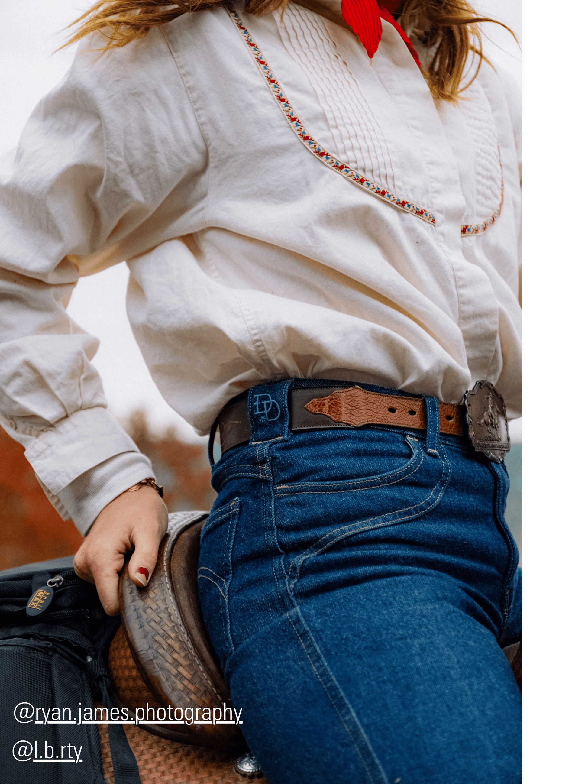 Person wearing a white shirt with embroidery, blue jeans, and a brown belt with a visible brand logo.