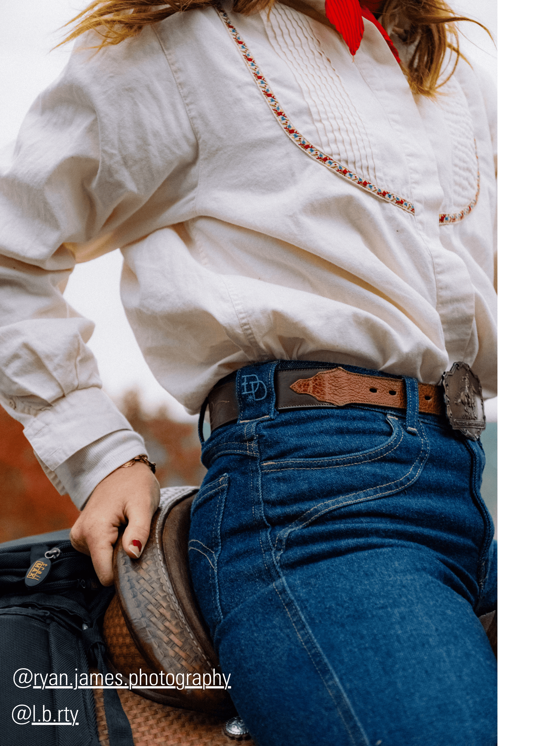 Person wearing a white blouse with embroidery and blue jeans, holding a horse saddle.