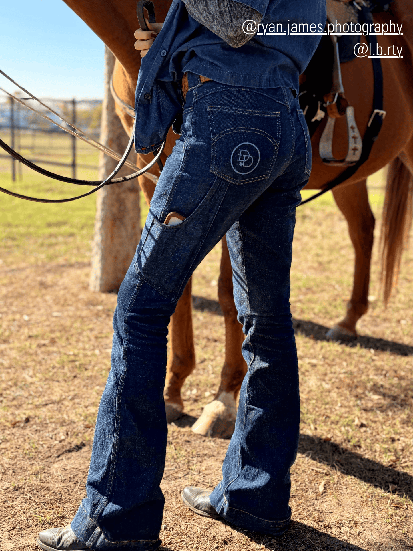 Person wearing blue jeans with a logo, standing next to a horse in an outdoor setting.